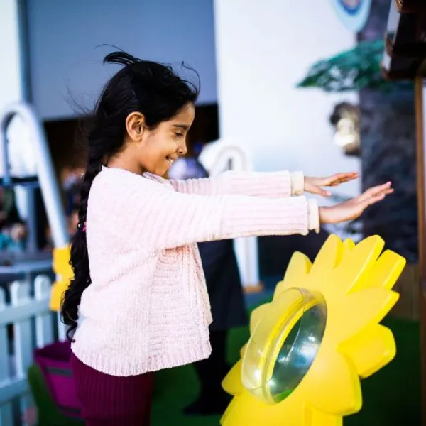 A child is stood in We The Curious science centre in a pink jumper. Stood Infront of a large, yellow, plastic flower and they are holding their hands up over it as it blows wind in their direction.