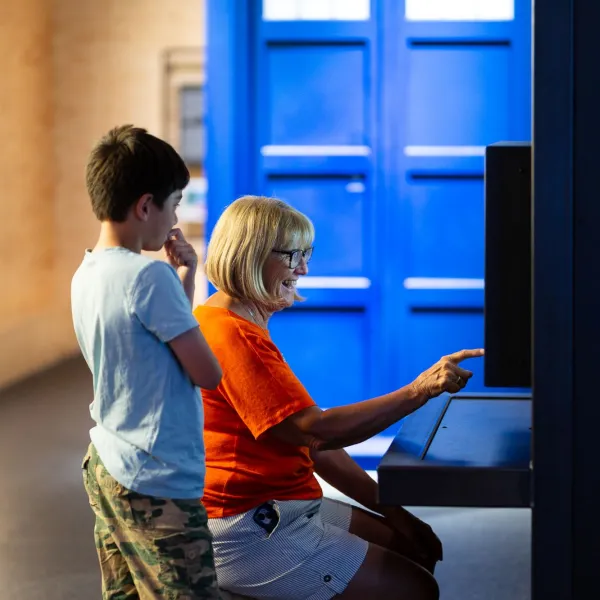 An adult and a child are in a science centre. The adult is sat down pointing at a screen and the child is stood next to them watching.